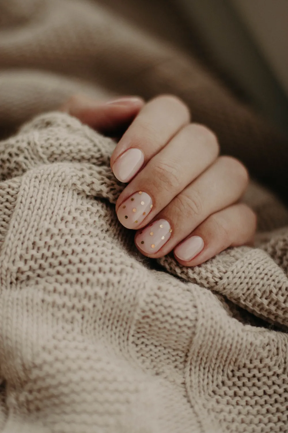 Neutral nude nails finished with tiny gold dots, resting on a knitted jumper.